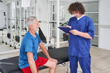 Senior man waiting for prescriptions on preventive examination of doctor chiropractor. Male manual therapist writing recipe to his male patient at rehabilitation centre
