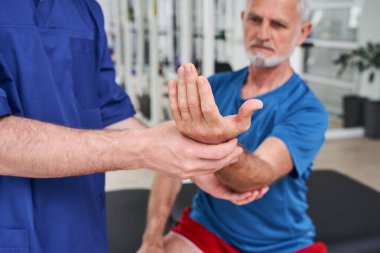 Senior man receiving massage by male physic therapist of his arm due to injury. Focus at the hand. Rehabilitation concept