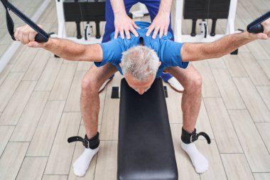 High angle view of the grey haired man working with young physiotherapist in rehabilitation centre. Retirement male stretching his back