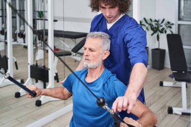 Focused man chiropractor or osteopath correcting old man patients movements during rehabilitation in manual therapy clinic. Trainer during work concept