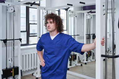 Young curly physical therapist standing at the rehabilitation centre while working with different equipment. Recovery and treatment concept