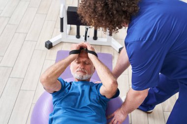 High angle view of the senior man laying on a fitness ball in physical rehabilitation therapy with his trainer. Male stretching his arms and muscles