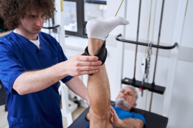 Osteopathy assistant stretching leg of his retirement patient with muscle pain for physiotherapy. Chiropractor treating senior man with injury, helping with physical recovery and remedy