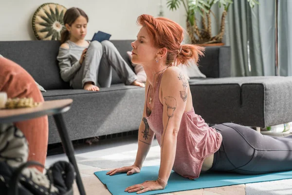 Ginger woman stretching at the cobra pose while her little daughter watching cartoons at the tablet at the sofa. Stock photo