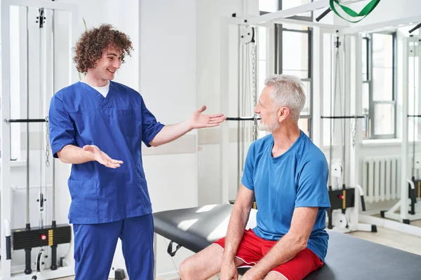 Cheerful Young Trainer Smiling Congratulating His Senior Patient Successful Treatment — Foto Stock