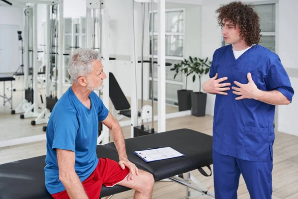 Physiotherapist talking to aged man with injury how to recover at clinic. Chiropractor giving advices to senior patient with back pain and muscle pressure for recovery