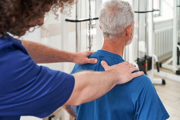 Back view of the male physiotherapist working on elderly mans tension his shoulders. Stretching and pressure release concept