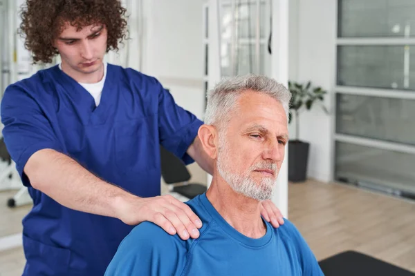 Medical specialist giving shoulder massage to senior man during the recovery session. Rehabilitation centre concept. Stock photo
