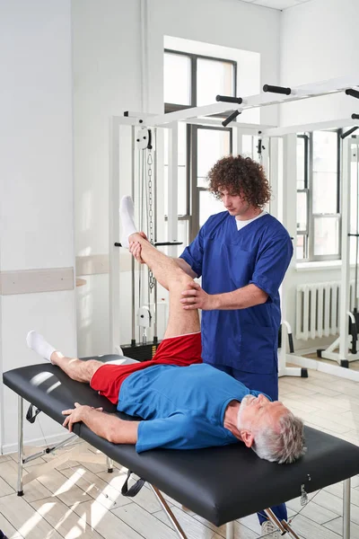 Physiotherapist doing healing treatment on mans leg. Massage therapist working with senior patient at medical office. Stock photo
