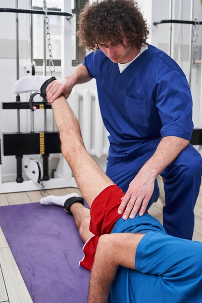 Senior male laying at his back at the yoga mat and stretching leg under assistance of young coach. Recovery at the retirement age concept. Stock photo