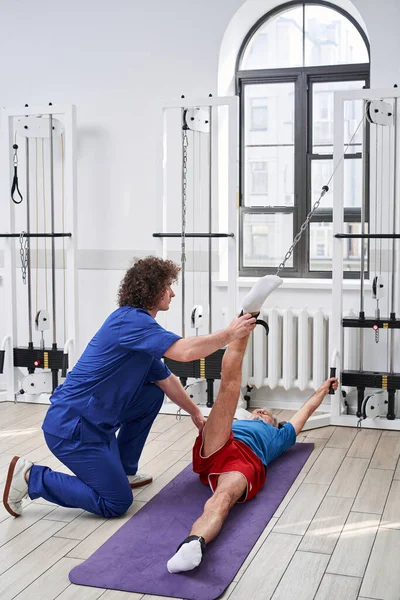 Vertical view of the physical therapy elderly patient using physiotherapy bands for rehabilitation of his legs. Young coach helping to him. Stock photo
