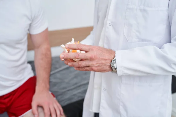 Cropped view of the doctor holding anatomical human bone model, during physiotherapy session with male patient. Physio treatment and kinesiotherapy concept