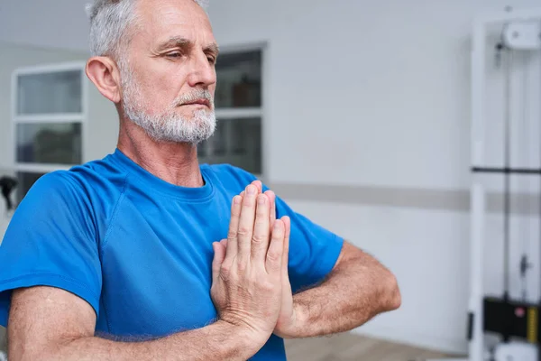 Focused old man training at the rehabilitation centre, trying yoga exercises, making prayer gesture. Recovery and physical therapy concept