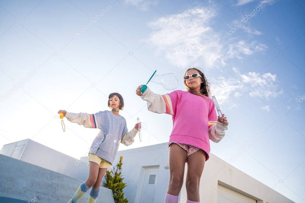 Little girlfriends resting together outdoors, blowing soap bubbles and having fun at roller skates. Summer and friendship concept