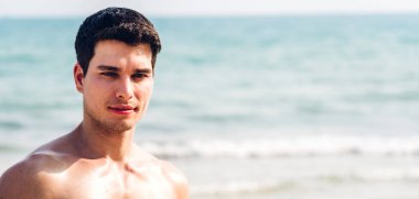 Gülümseyen yakışıklı seksi adamın portresi kaslı fit vücudunu gösteriyor tropik sahilde durup eğleniyor ve rahatlıyor.