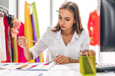 Genç ve güzel sahibinin portresi iş kadını moda tasarımcısı şık oturma ve çalışma. Genç tasarımcı kız, moda tasarım atölyesinde masaüstü ürünü ve renkli kumaşlar kullanıyor.
