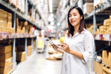 Gülümseyen Asyalı mühendis kadının portresi, malzeme ve malzemelerin depo geçmişine sahip raflarda kontrol edilmesi için tablet bilgisayardan ayrıntılı talimat vermektedir. Lojistik ve ticari ihracat