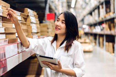 Gülümseyen Asyalı mühendis kadının portresi, malzeme ve malzemelerin depo geçmişine sahip raflarda kontrol edilmesi için tablet bilgisayardan ayrıntılı talimat vermektedir. Lojistik ve ticari ihracat