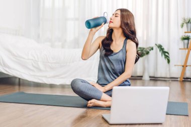 Spor kıyafetli, spor elbiseli, Asyalı, sıska bir kadının portresi. Evde oturmuş, bir şişeden su içiyor. Yatak odasında dizüstü bilgisayarla spor yapıyor.