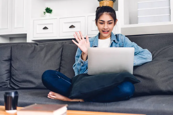 Genç gülümseyen Asyalı kadın dizüstü bilgisayar kullanıyor ve evdeki iş arkadaşlarıyla çevrimiçi toplantı yapıyor. Genç yaratıcı kız kulaklıkla video konuşması yapıyor. Ev konseptinden çalışma.