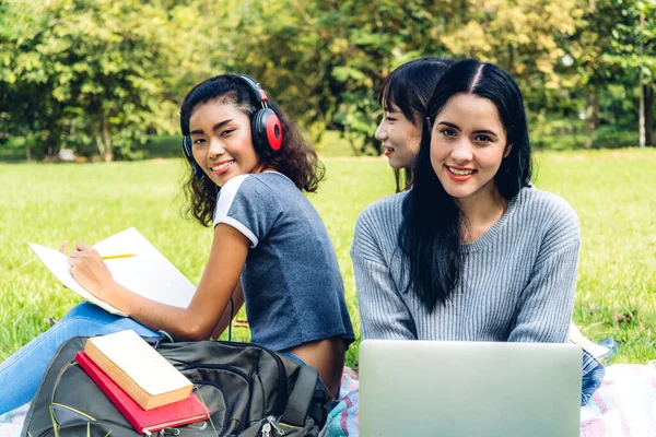 Bir grup gülümseyen uluslararası öğrenci ya da gençler dizüstü bilgisayarlarıyla oturup öğreniyorlar ödevlerini üniversitede parkta birlikte okuyorlar. Eğitim ve arkadaşlık kavramı.