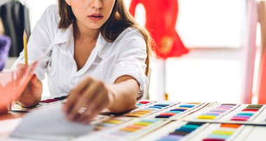 Genç ve güzel sahibinin portresi iş kadını moda tasarımcısı şık bir şekilde oturuyor ve çalışıyor. Genç tasarımcı kız, moda atölyesinde masa üstü ürünü ve renkli kumaşlar kullanıyor.
