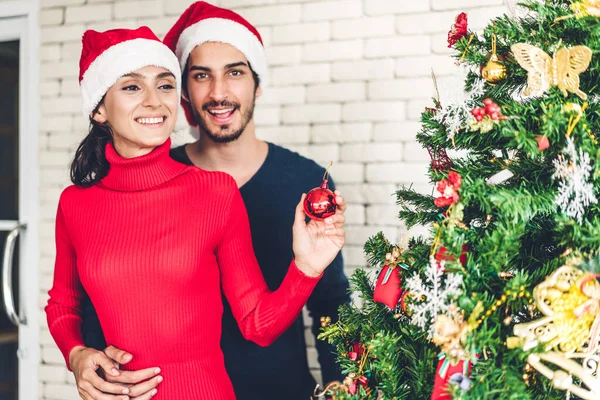 Romantic sweet couple in santa hats having fun decorating christmas tree and smiling while celebrating new year eve and enjoying spending time together in christmas time at home