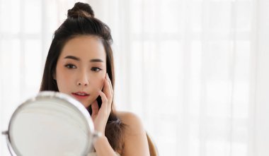 Genç, güzel Asyalı bir kadının, temiz, sağlıklı, beyaz bir tenle aynaya bakması. Asyalı bir kızın elleriyle yüzüne dokunması ve evde krem sürmesi. Spa ve güzellik konsepti.