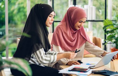 Gülümseyen mutlu Asyalı güzel Müslüman kadın dizüstü bilgisayarla rahatlıyor, çalışıyor ve analiz ediyor. Başlangıç projesi planı ve evdeki beyin fırtınası ile strateji tartışıyorlar.