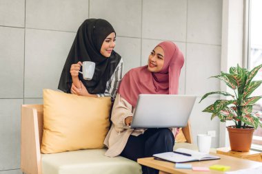 Gülümseyen mutlu Asyalı güzel Müslüman kadın dizüstü bilgisayarla rahatlıyor, çalışıyor ve analiz ediyor. Başlangıç projesi planı ve evdeki beyin fırtınası ile strateji tartışıyorlar.