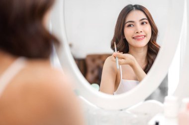 Güzel, güzel Asyalı bir kadının, temiz, sağlıklı, beyaz tenli, aynaya bakan gülümsemesi. Elinde makyaj fırçası olan Asyalı bir kız. Yüzüne makyaj malzemesi konmuş.