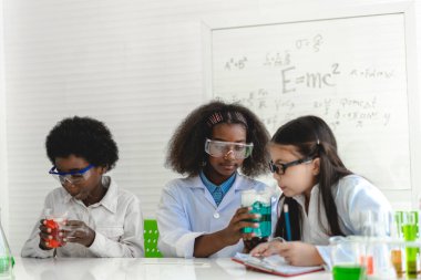 Bir grup genç öğrenci kimyasal bir deney yapıyor ve deney tüpünü el ele tutuyorlar. Laboratuvar sınıfında, okuldaki masada. Eğitim konsepti.