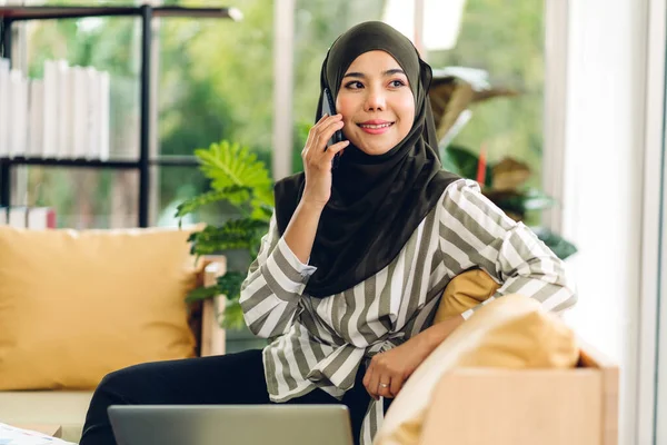 Genç gülümseyen mutlu Asyalı Müslüman kadın dizüstü bilgisayarını kullanarak rahatlıyor ve evde başlangıç projesi planıyla strateji analizi yapıyor.