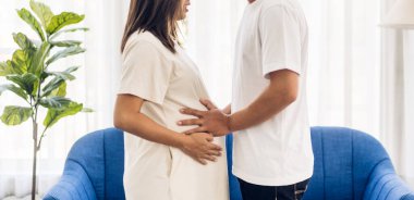 Romantik tatlı Asyalı koca kucaklıyor ve gülümsüyor. Asyalı hamile kadın. Hamilelik bekleyen adam karnında bebeğini evde tutuyor.