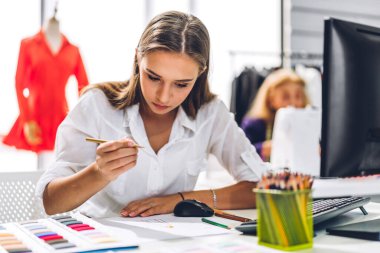 Genç ve güzel sahibinin portresi iş kadını moda tasarımcısı şık bir şekilde oturuyor ve çalışıyor. Genç tasarımcı kız, moda atölyesinde masa üstü ürünü ve renkli kumaşlar kullanıyor.
