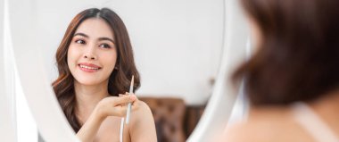 Güzel, güzel Asyalı bir kadının, temiz, sağlıklı, beyaz tenli, aynaya bakan gülümsemesi. Elinde makyaj fırçası olan Asyalı bir kız. Yüzüne makyaj malzemesi konmuş.