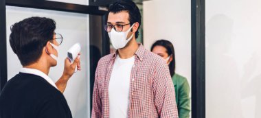 Bir grup profesyonel Asya işi ve ofiste koruyucu maske takarak koronavirüs için karantinada çalışmadan önce vücut ısısını ölçmek için kızılötesi termometre kullanılıyor.