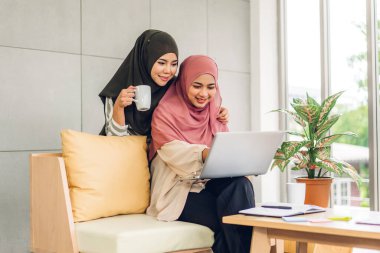 Gülümseyen mutlu Asyalı güzel Müslüman kadın dizüstü bilgisayarla rahatlıyor, çalışıyor ve analiz ediyor. Başlangıç projesi planı ve evdeki beyin fırtınası ile strateji tartışıyorlar.