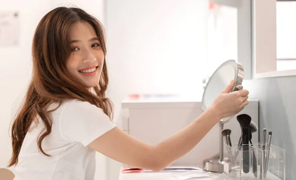 Smiling of young beautiful pretty asian woman clean fresh healthy white skin looking at mirror.asian girl touching on her face with hand and applying cream at home.spa and beauty concept