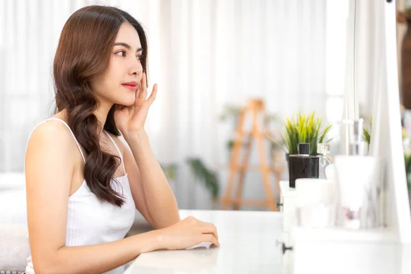 Smiling of young beautiful pretty asian woman clean fresh healthy white skin looking at mirror.asian girl touching on her face with hand and applying cream at home.spa and beauty concept