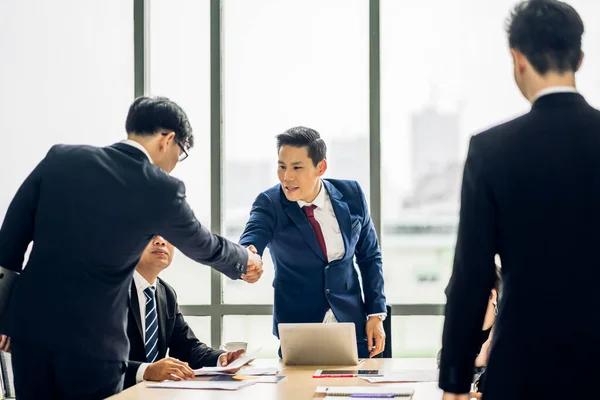 在现代办公室里 两个穿着雅致西装的亚洲商业伙伴在一群随意拍手的同事面前成功地握手 — 图库照片