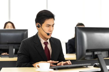 Mutlu çağrı merkezi gülen Asyalı iş adamı müşteri, çağrı merkezindeki masaüstü bilgisayarında çalışan ve kulaklıkla konuşan ekip telefon hizmetlerini desteklemeye yardım eder.