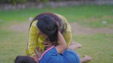 Indian Mother with her cute son, child playing together, throwing grasses on each other, laughing, hugging, falling on ground  having fun, joy  in beautiful green garden. Mother's Day concept.