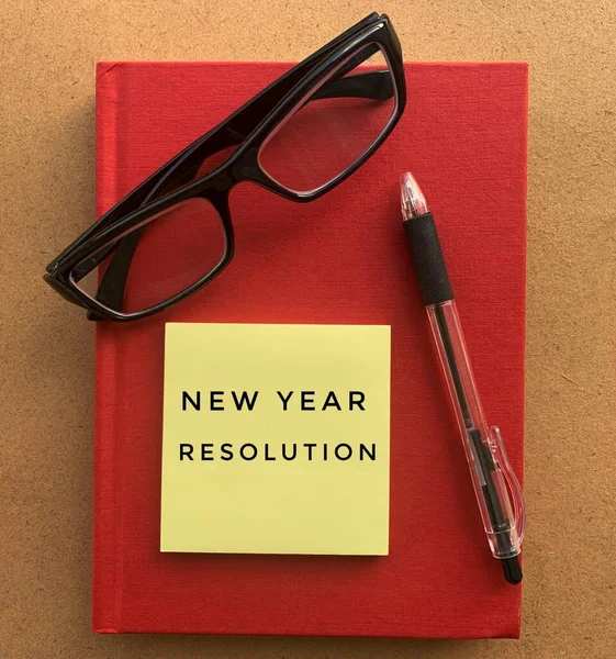 Tekst noworocznej rezolucji na lepkiej nucie z napisem, szkłem i czerwonym tłem książki — Zdjęcie stockowe