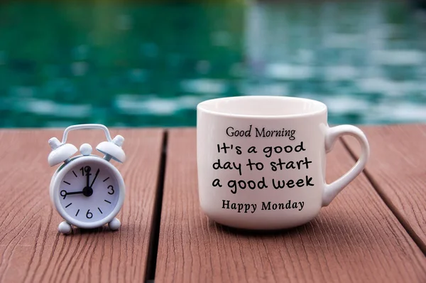 Белый будильник на деревянном полу с утренним текстом цитаты на чашке кофе - Это хороший день, чтобы начать хорошую неделю. С размытым плавательным фоном. — стоковое фото