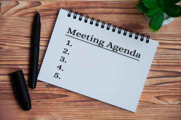 Meeting agenda text on notepad with plant, pen and wooden table background. Meeting concept