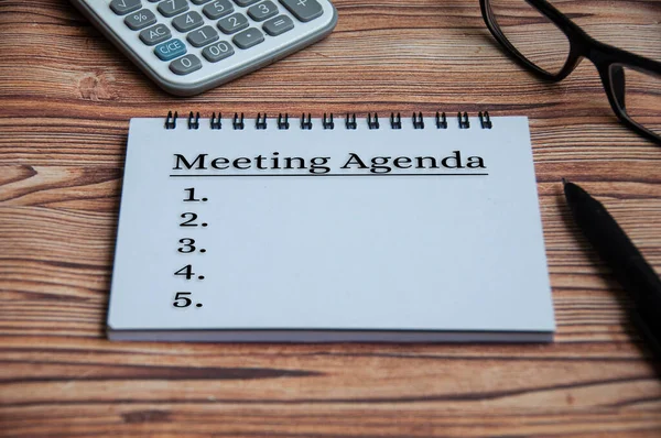 Meeting agenda text on notepad with calculator, pen, glasses and wooden table background. Meeting concept