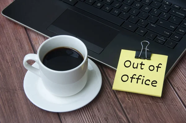 Coffee cup and laptop on wooden table with yellow notepad with text - Out of office. Business concept