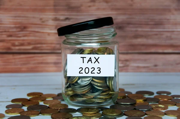 Tax 2023 label on coin jar with scattered coins on wooden table. Tax concept