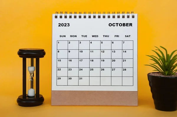 October 2023 desk calendar with minutes glass and plant on yellow background. Calendar concept